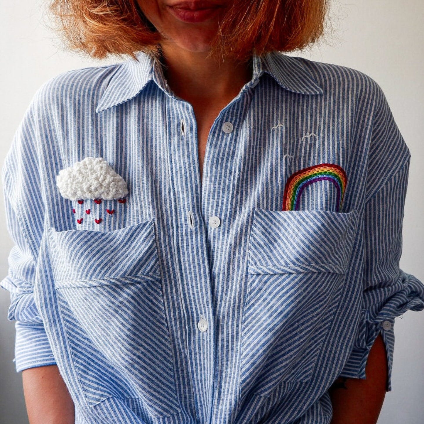 Blue striped shirt with embroidered cloud and rainbow designs on a plain background