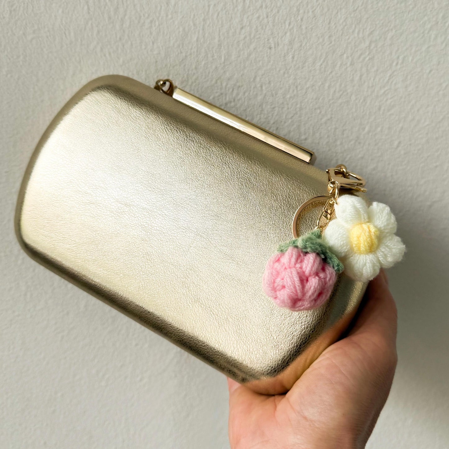 Gold clutch with a floral keychain held by a hand against a neutral background