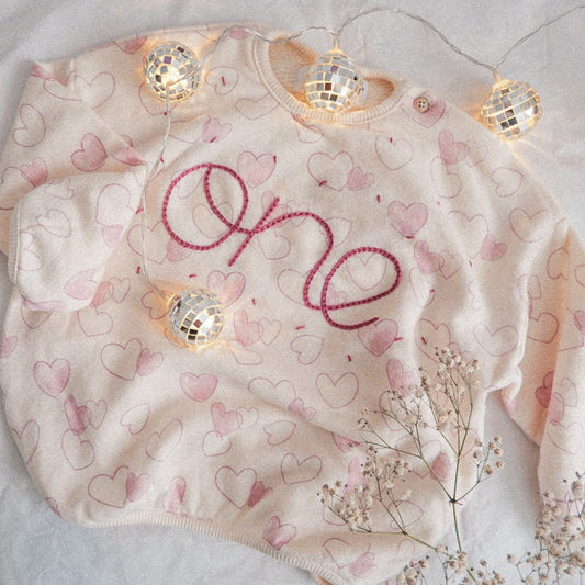 Baby romper with heart pattern and 'one' embroidery, surrounded by decorative lights and flowers on a light background.