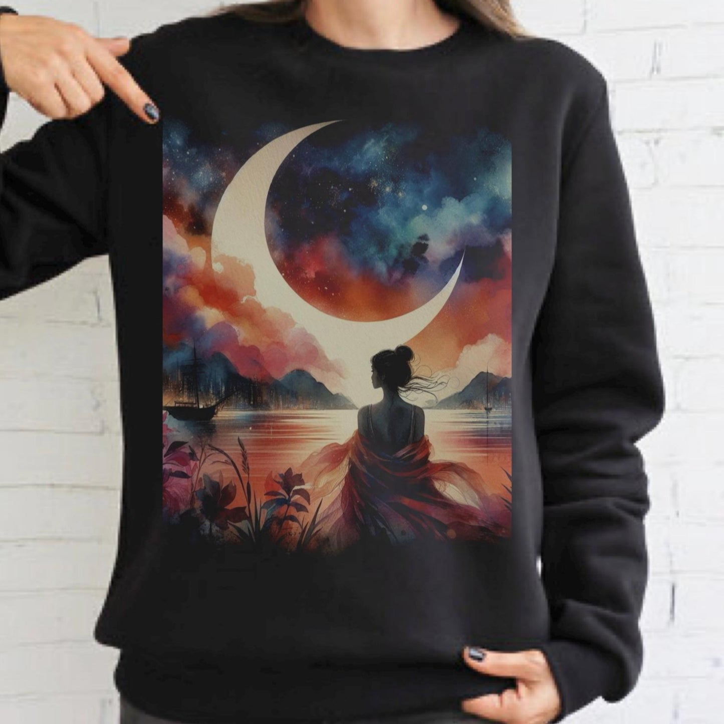 Black sweatshirt with a colorful design of a woman sitting by a lake with a crescent moon above, held up against a white brick wall.