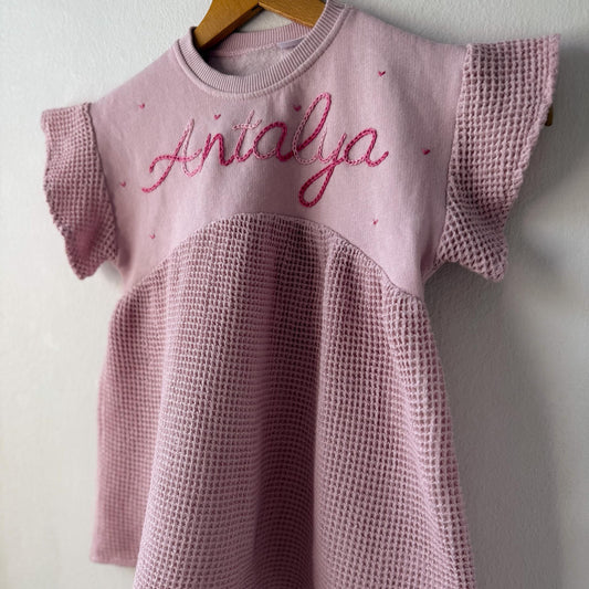 Pink textured dress with 'Antalya' embroidery on a wooden hanger against a white wall.