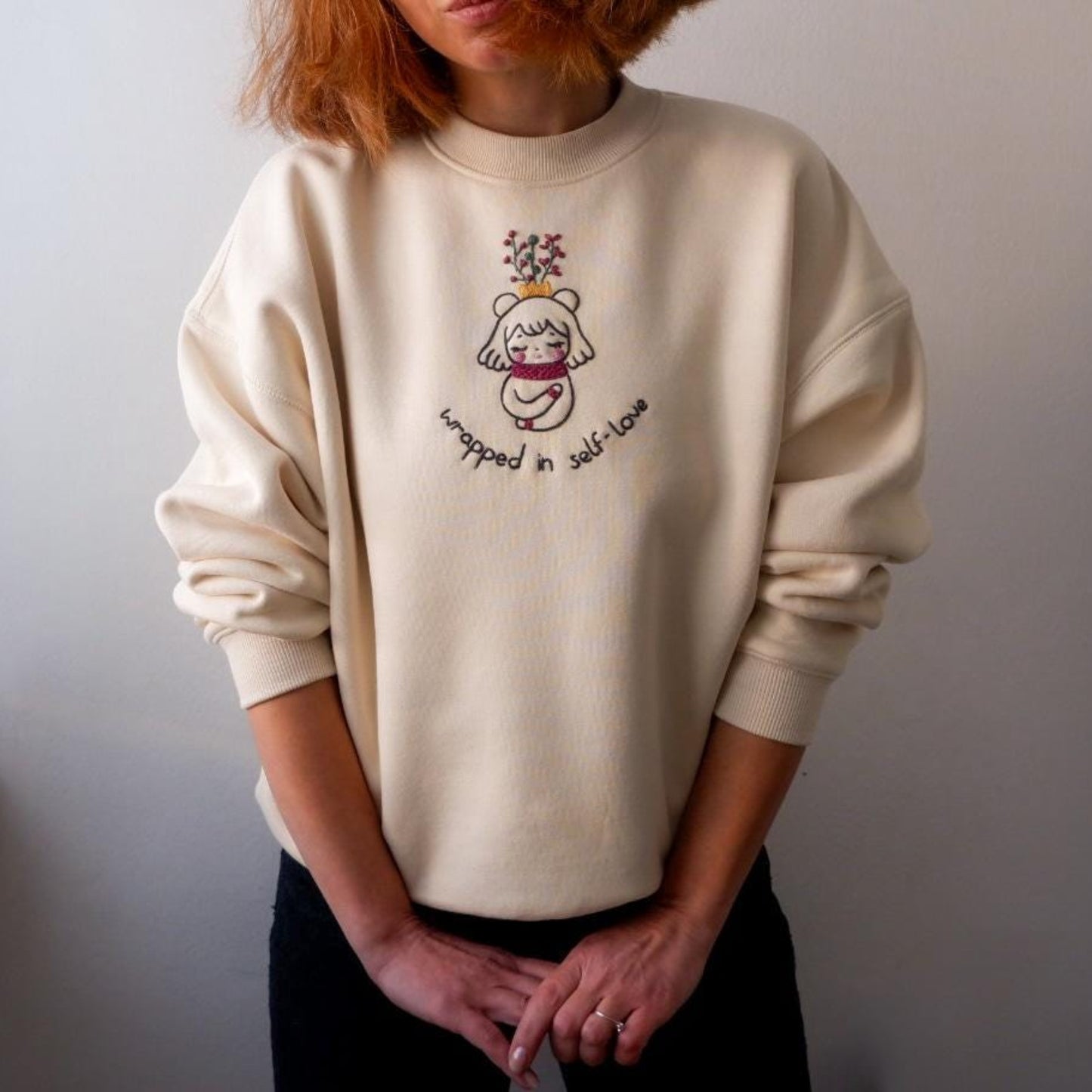 Person wearing a cream sweatshirt with a graphic design and text against a plain background