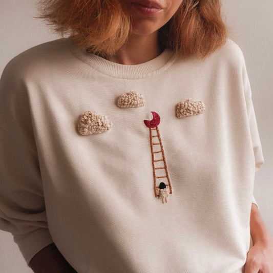 Person wearing a beige sweater with embroidered cloud and ladder design.