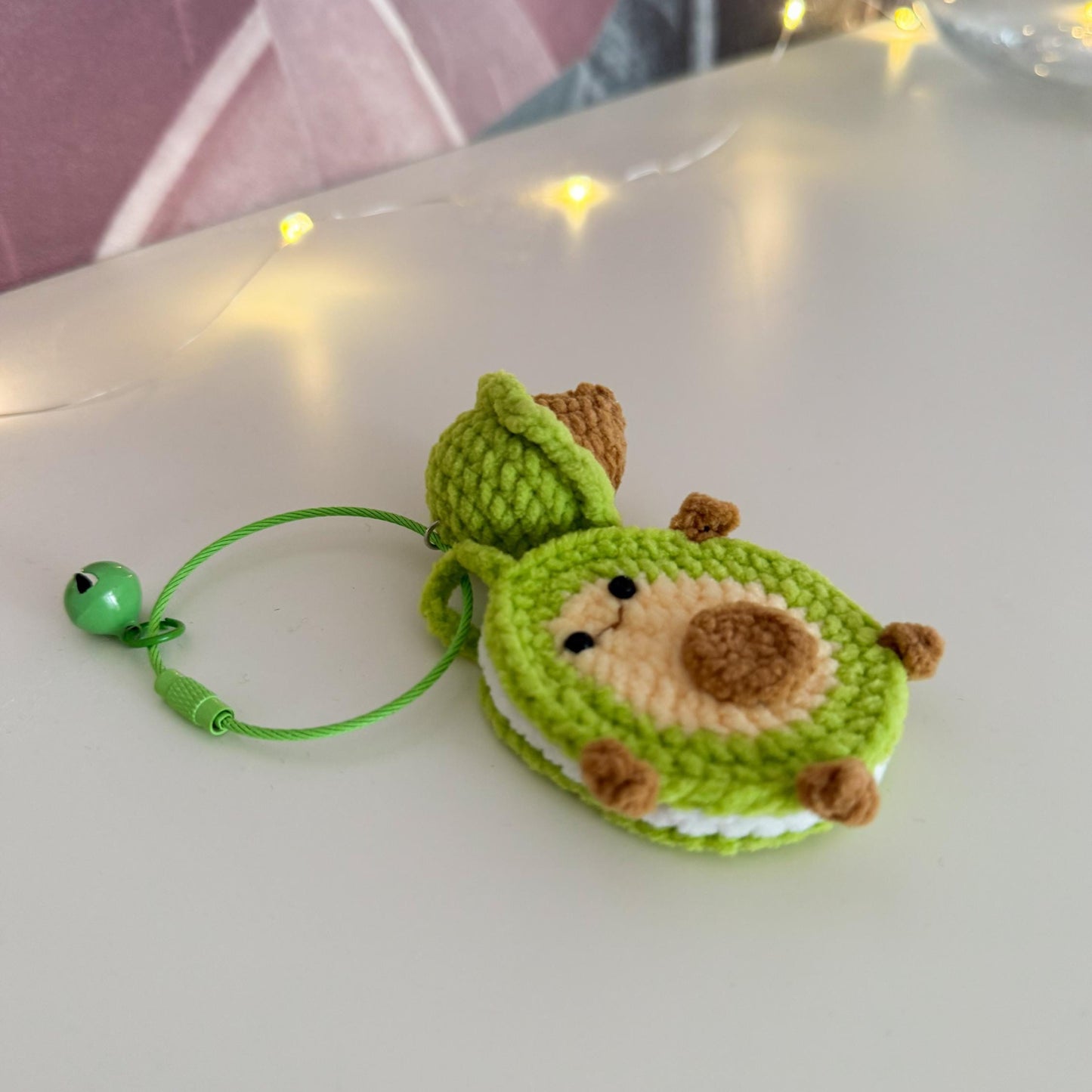 Green and brown crocheted keychain on a white surface with blurred lights in the background