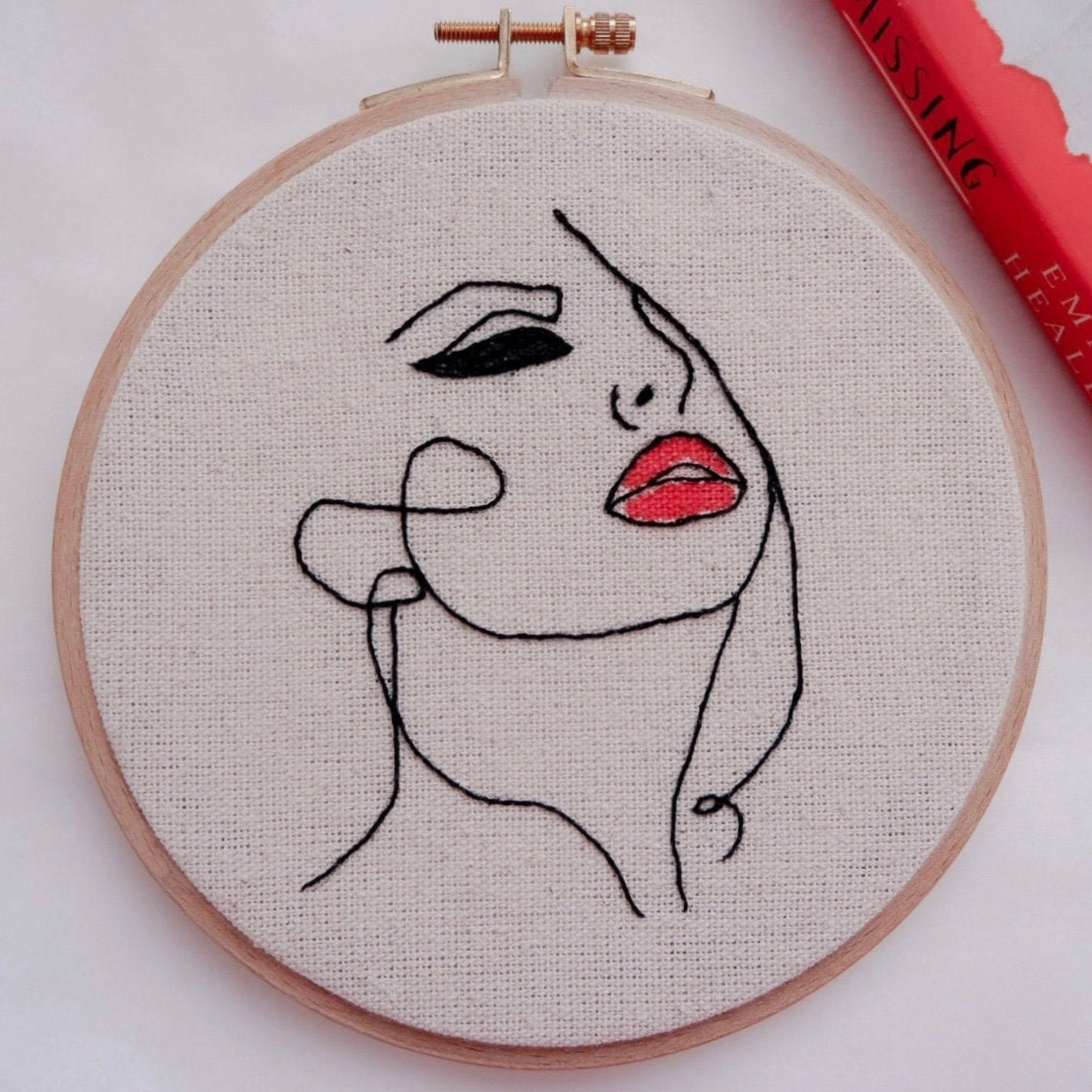 Hand embroidery hoop art featuring a minimalist line drawing of a woman's face with red lipstick, primarily in black and red colors on a beige background.