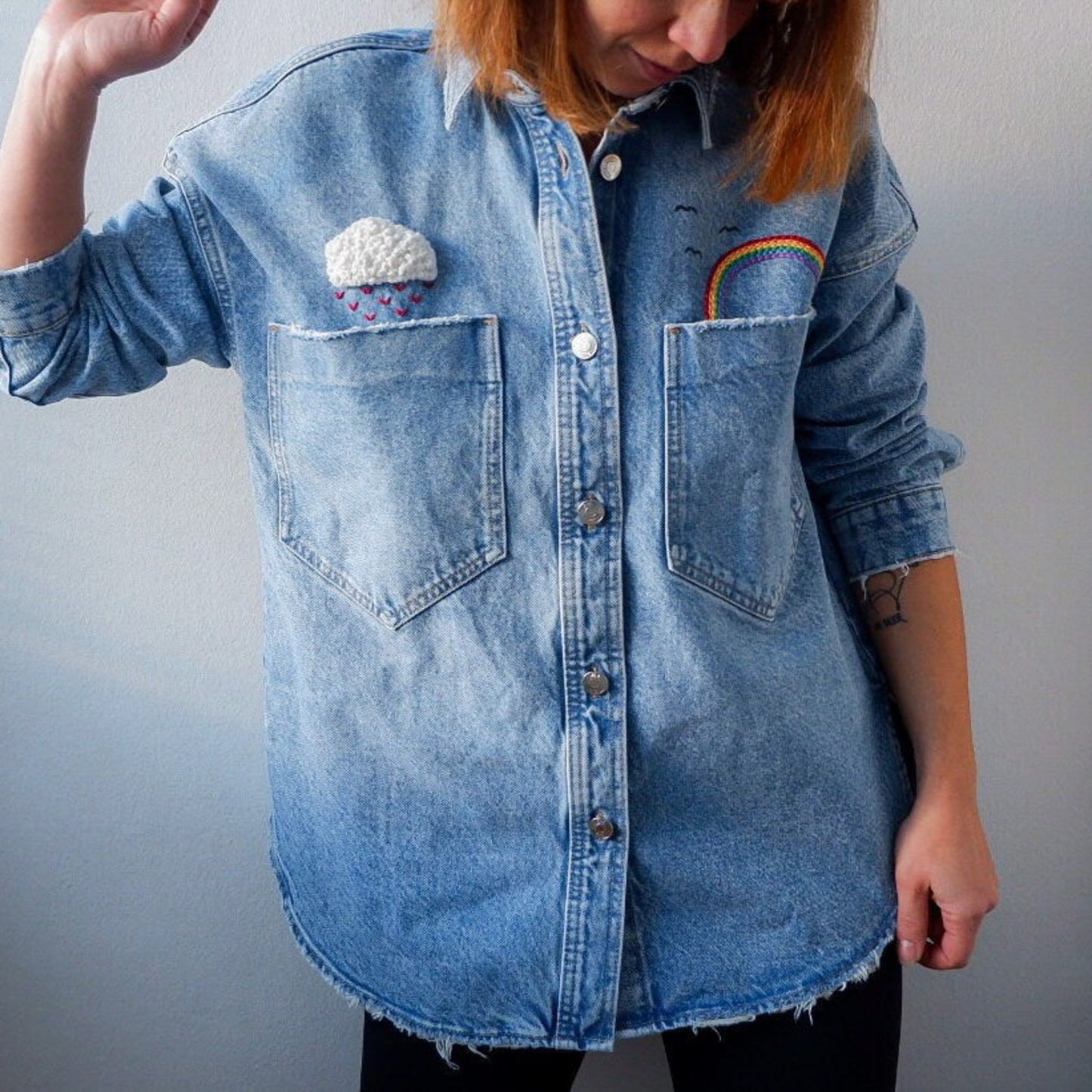 Person wearing a denim shirt with embroidered cloud and rainbow design on a plain background