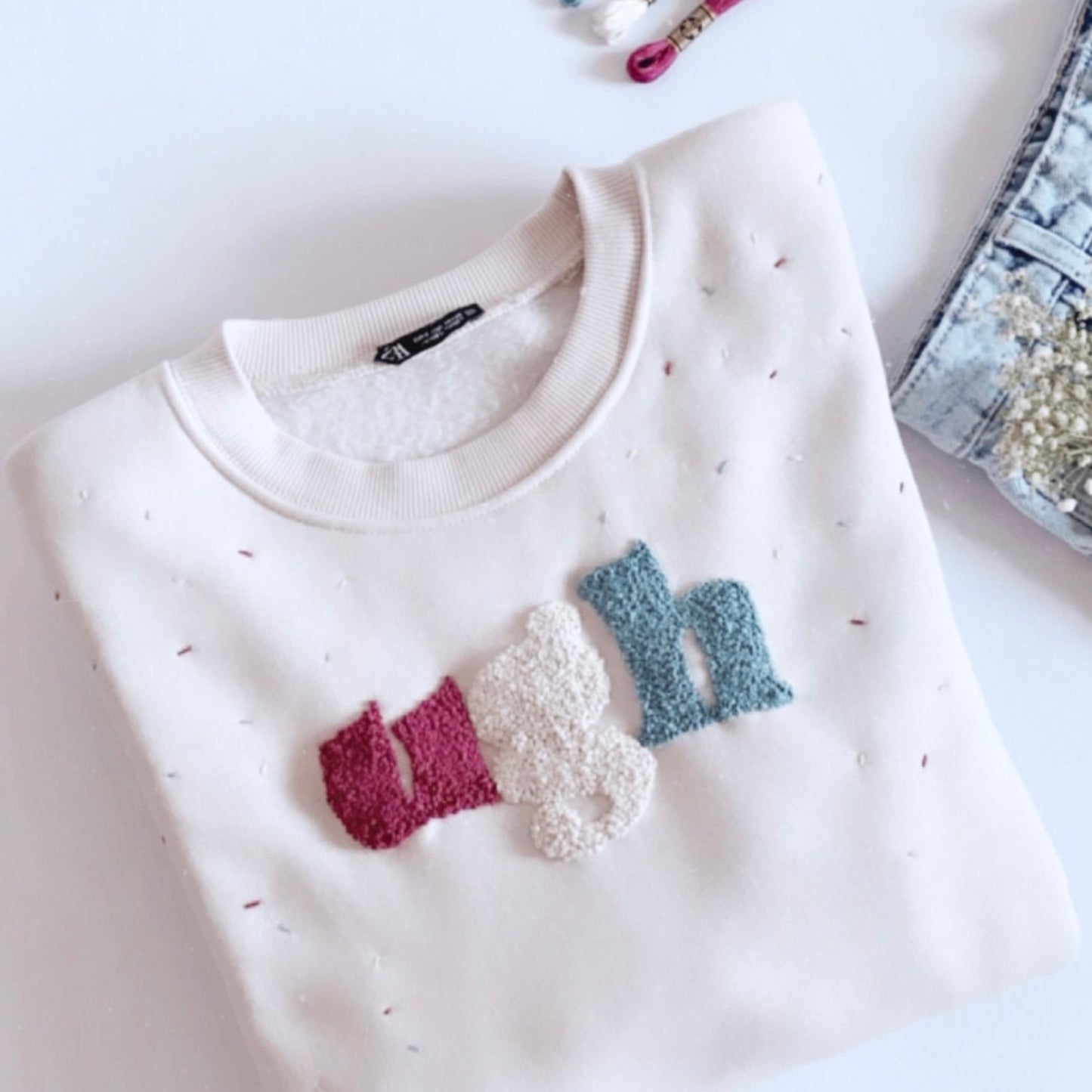 A white sweatshirt with 'ugh' hand embroidered in multicolored threads on the front.