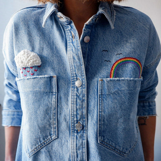 Blue denim shirt with embroidered designs on the pockets against a neutral background