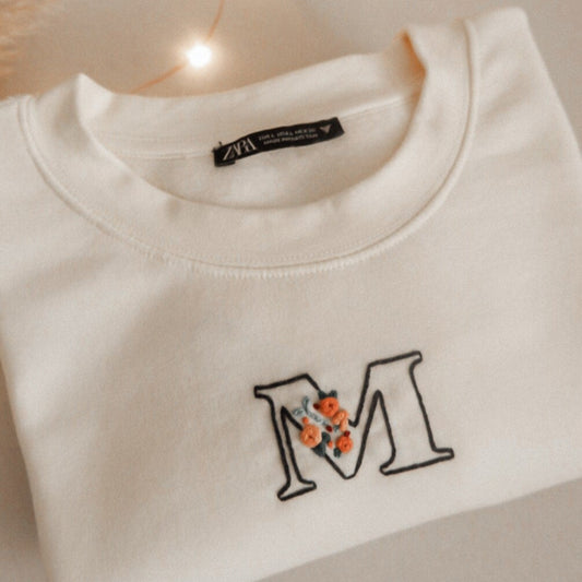 White sweatshirt with letter 'M' and floral design, placed on a light surface with a candle and string lights in the background.