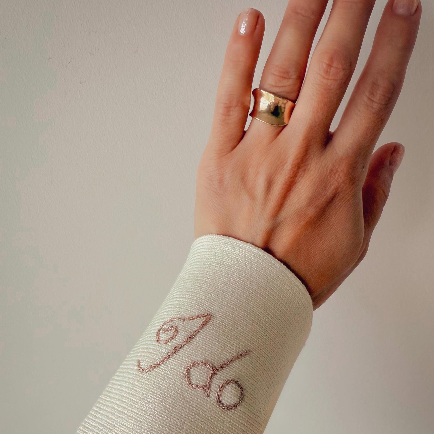 Hand wearing a gold ring with a white sleeve featuring embroidered text on a plain background