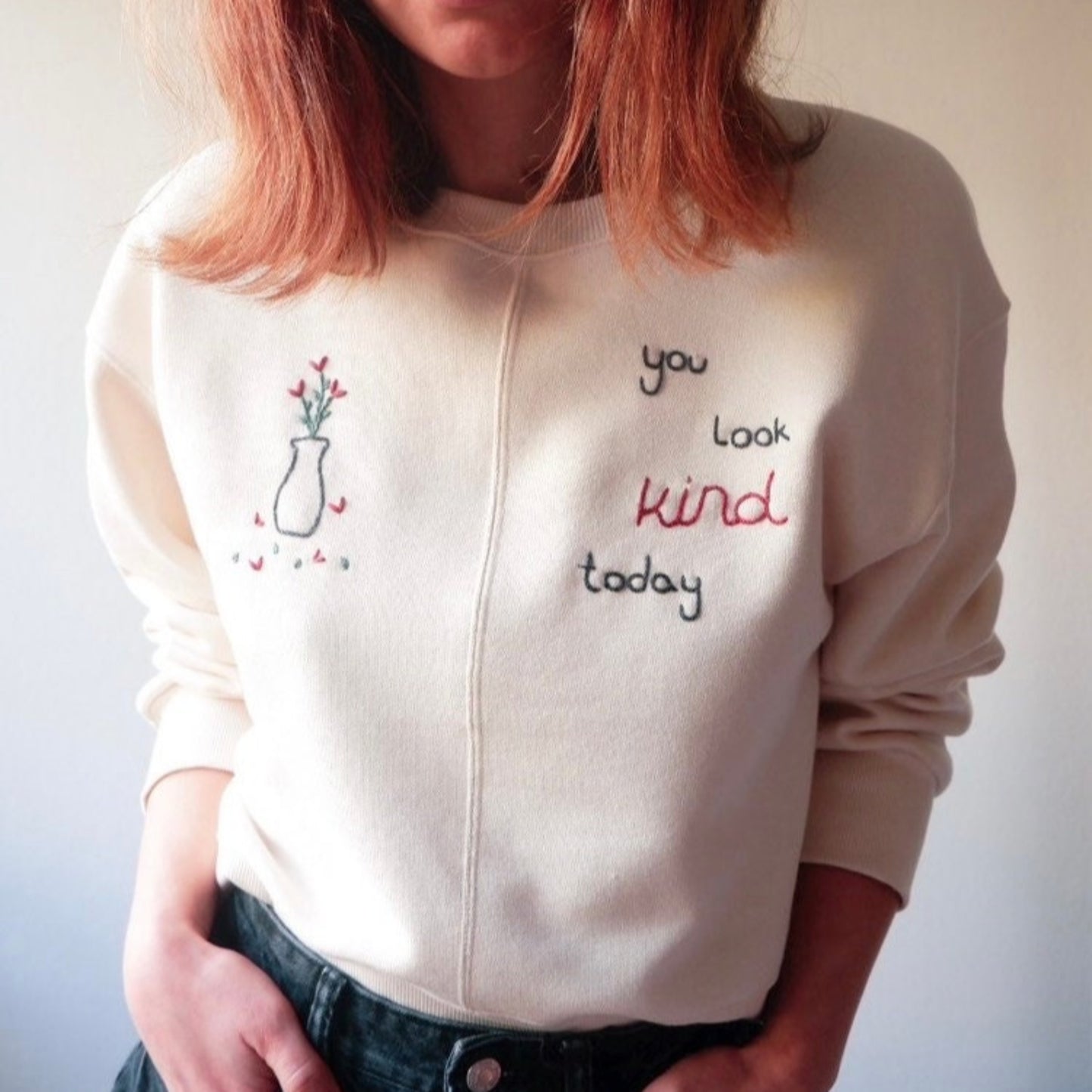 Person wearing a cream-colored sweatshirt with embroidered text and a vase design.