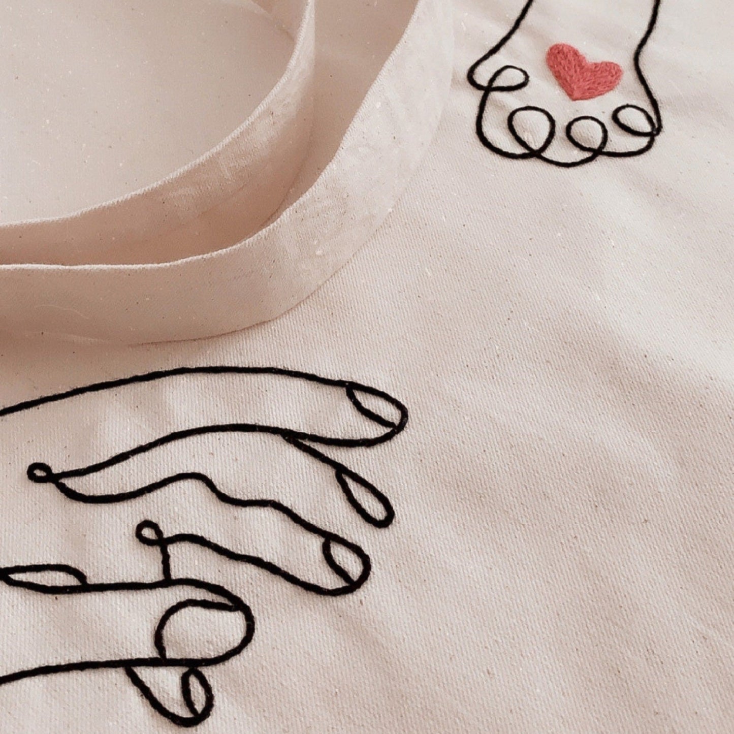 Beige fabric with black line art of hands holding a heart and a red heart embroidered on it.