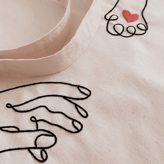 Beige fabric with black line art of hands holding a heart and a red heart embroidered on it.