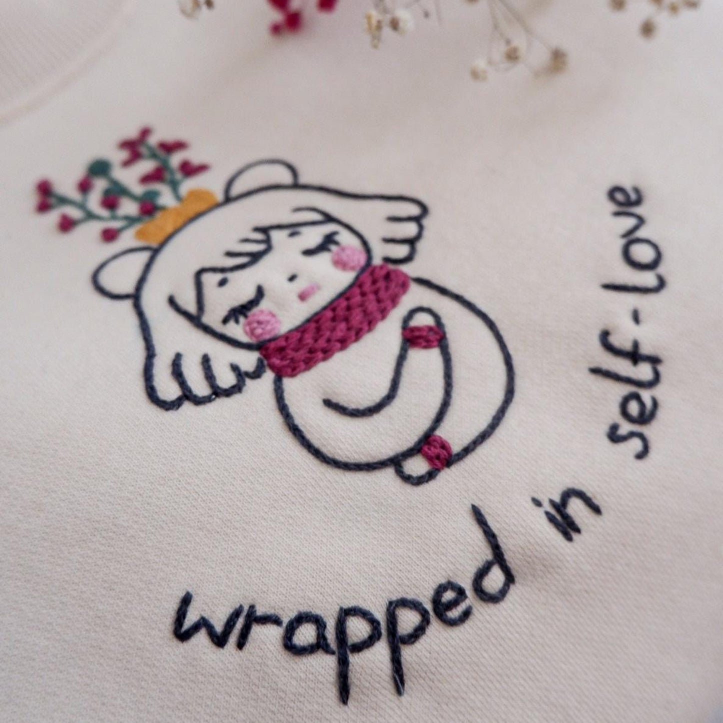 Embroidered design on fabric with a cartoon character and text 'wrapped in self-love'.