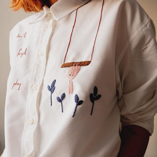 White shirt with embroidered design of a child on a swing and plants.