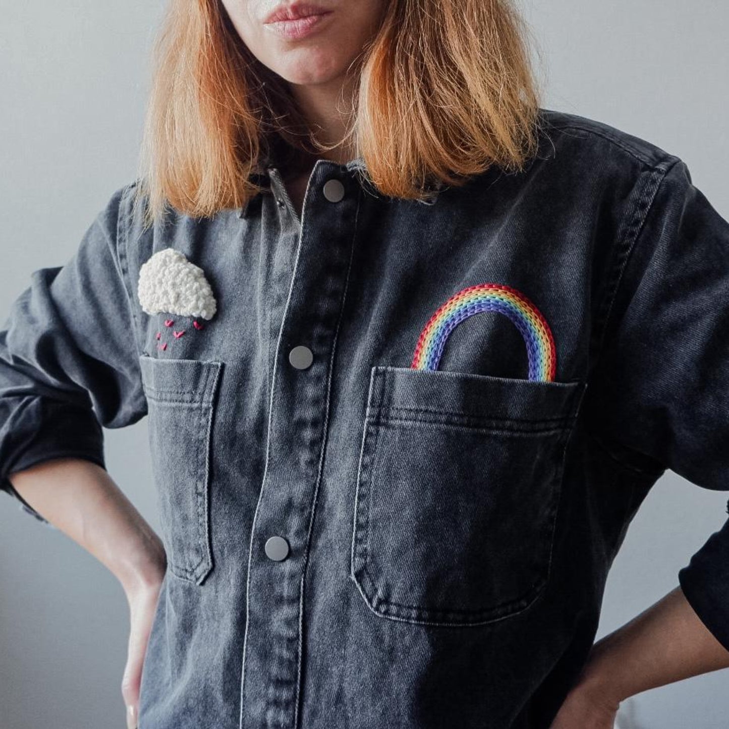 Person wearing a denim shirt with embroidered cloud and rainbow designs on a plain background