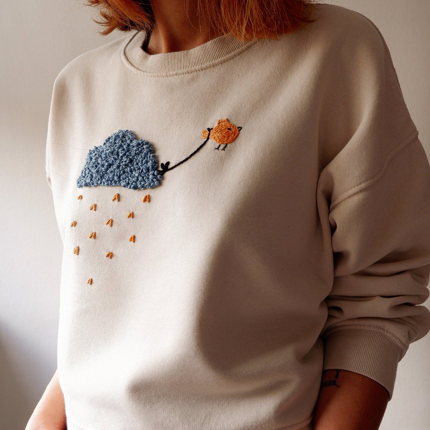 A beige sweatshirt with hand embroidered bird and cloud patterns on the front.