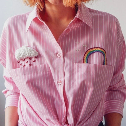 Person wearing a pink striped shirt with embroidered cloud and rainbow designs on the chest pocket.