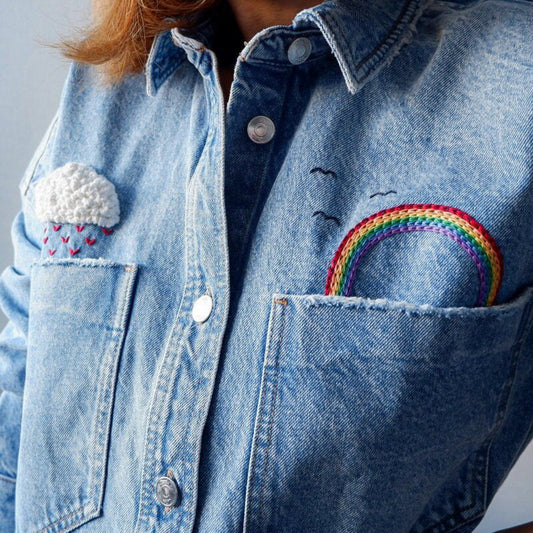Blue denim jacket with embroidered designs of a rainbow and cloud.