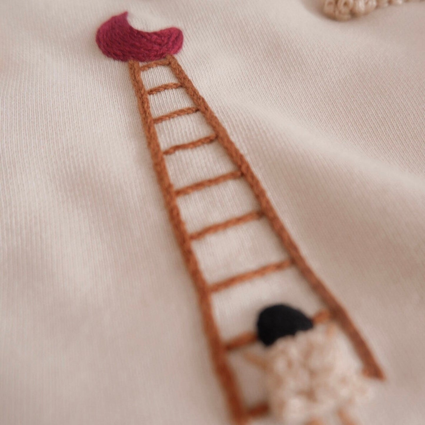 Embroidered design of a ladder with a girl at the bottom on a beige fabric background.