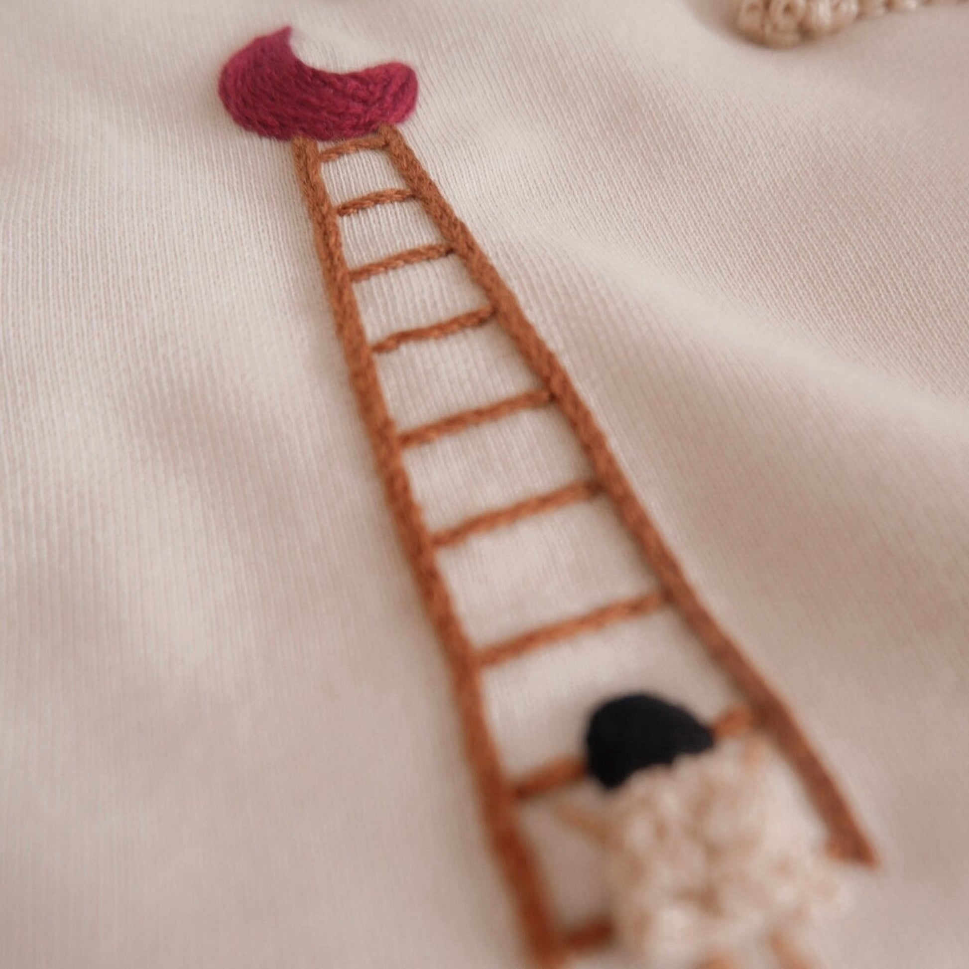 Embroidered design of a ladder with a girl at the bottom on a beige fabric background.