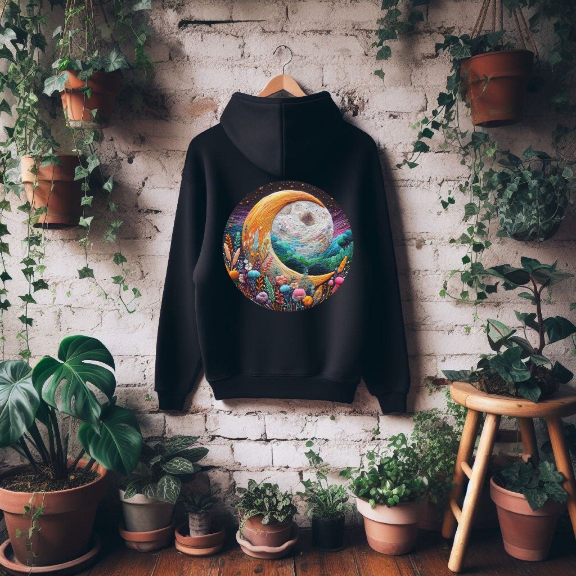 Black hoodie with a colorful design on a hanger against a brick wall with plants.