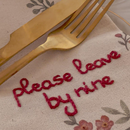 Napkin with 'Please leave by nine' embroidery, gold fork, and knife on floral patterned surface.