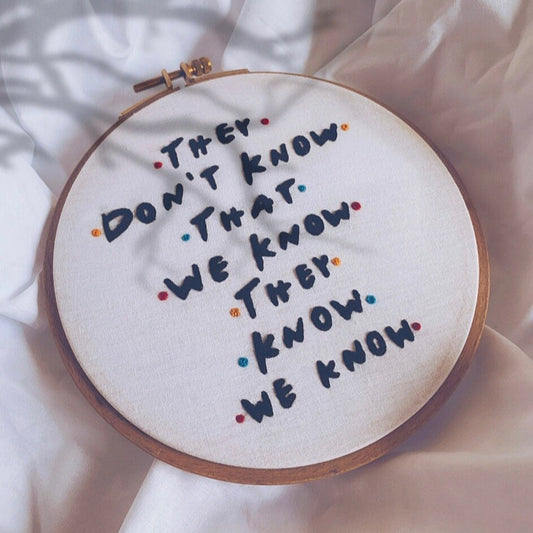 Embroidery hoop with text 'They don't know that we know, they know we know' on a white fabric background.