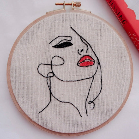 Hand embroidery hoop art featuring a minimalist line drawing of a woman's face with red lipstick, primarily in black and red colors on a beige background.