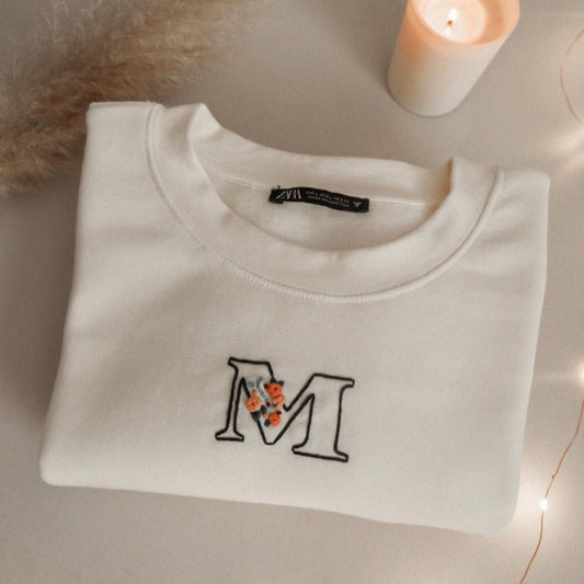 White sweatshirt with 'M' design on a neutral background with a candle and decorative elements.