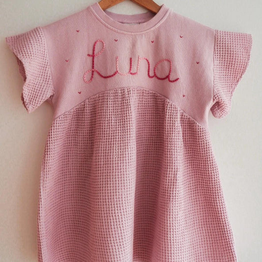 Pink checkered dress with 'Lina' embroidery on a wooden hanger against a white wall.
