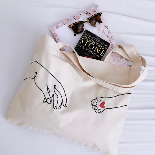 Beige tote bag with hand-drawn designs, book, and sunglasses on a white background