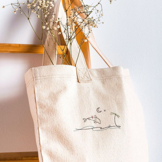 A beige canvas tote bag with hand embroidered sea and palm tree designs, displayed alongside some dried flowers.