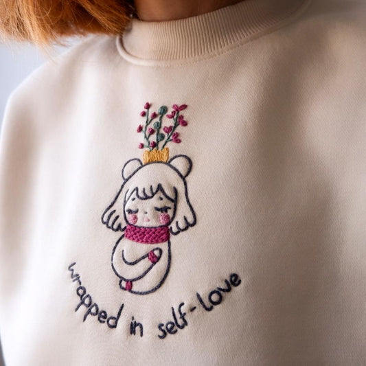 Beige sweatshirt with embroidered design of a girl holding flowers and text 'wrapped in self-love'.