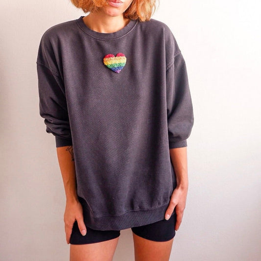 A person wearing a gray sweatshirt with a rainbow heart embroidered on the chest.