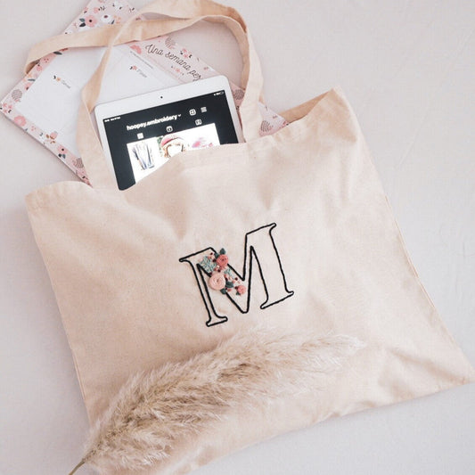 A beige cotton tote bag with personalized hand embroidery, displayed alongside a tablet and a partially visible floral patterned item, possibly a scarf.
