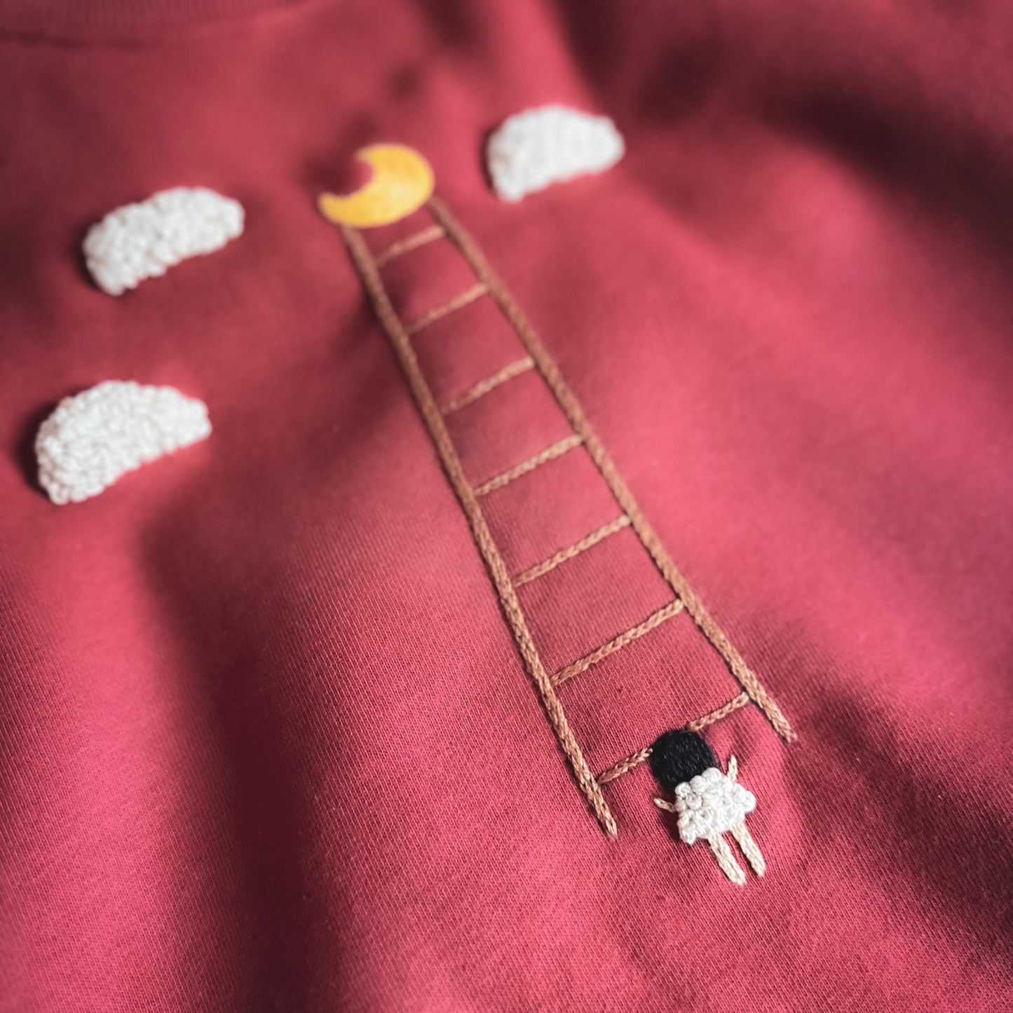 Red sweatshirt with embroidered ladder, moon, clouds, and figure design.